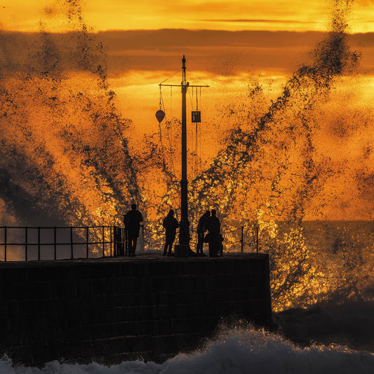 An Evocative Cornwall greetings card depicting people silhouetted at the end of the pier with a large wave crashing in front of them, lit with orange light by the setting sun