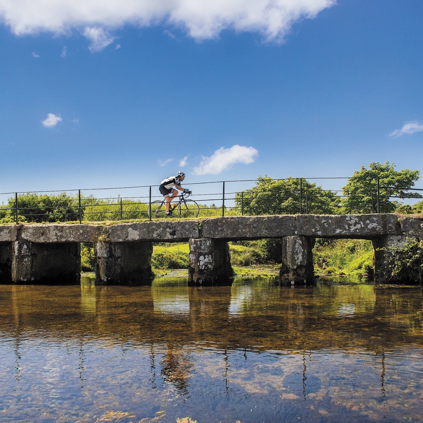 An Evocative Cornwall greetings card showing a cyclist crossing a granite clapper bridge on Bodmin Moor with still reflective water beneath and blue sky overhead