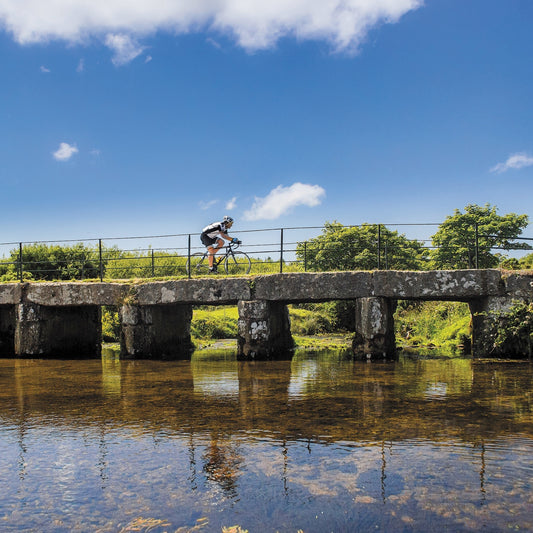 An Evocative Cornwall greetings card showing a cyclist crossing a granite clapper bridge on Bodmin Moor with still reflective water beneath and blue sky overhead