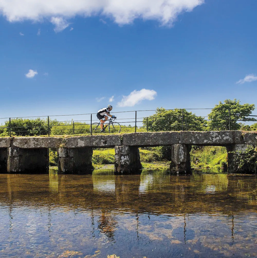 An Evocative Cornwall greetings card showing a cyclist crossing a granite clapper bridge on Bodmin Moor with still reflective water beneath and blue sky overhead