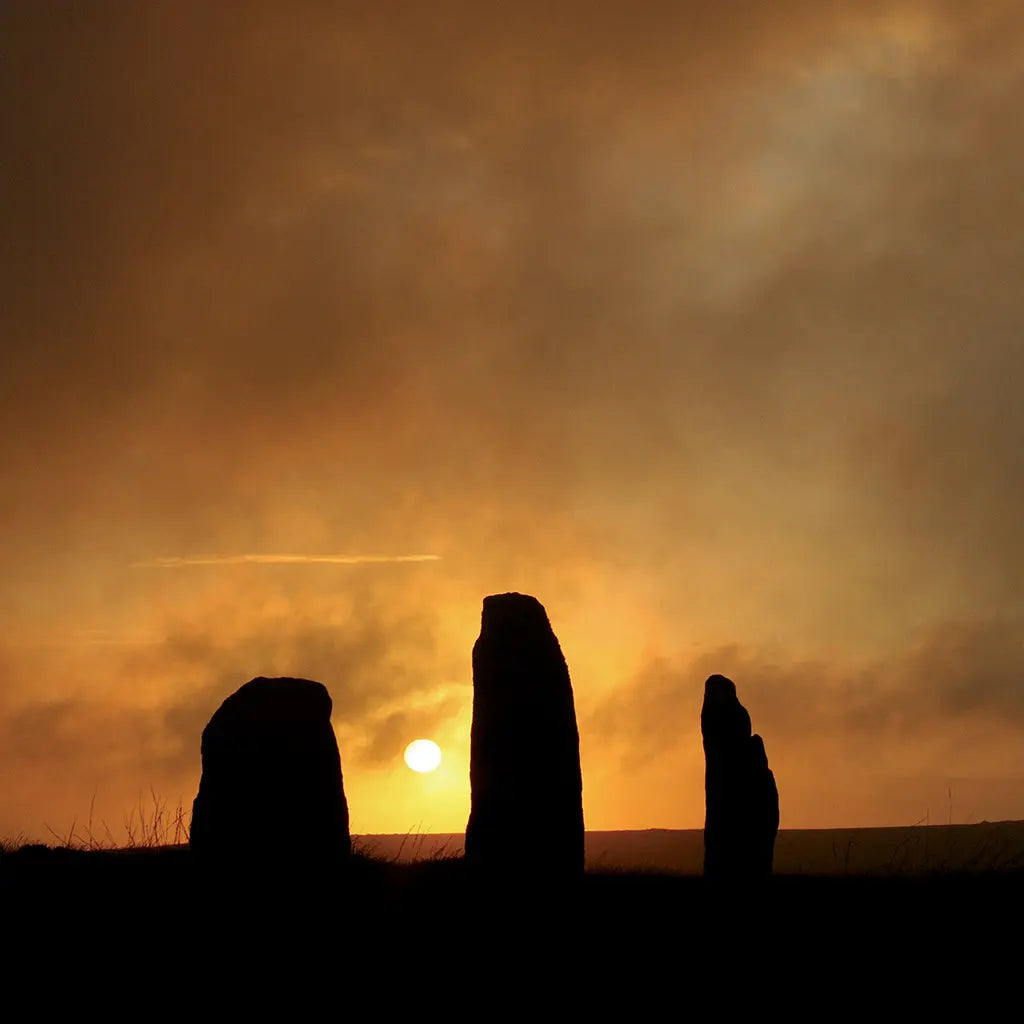 Cornish greetings card image of the sun setting between the stones of Nine Maidens stone circle in West Penwith, dramatic in black and orange