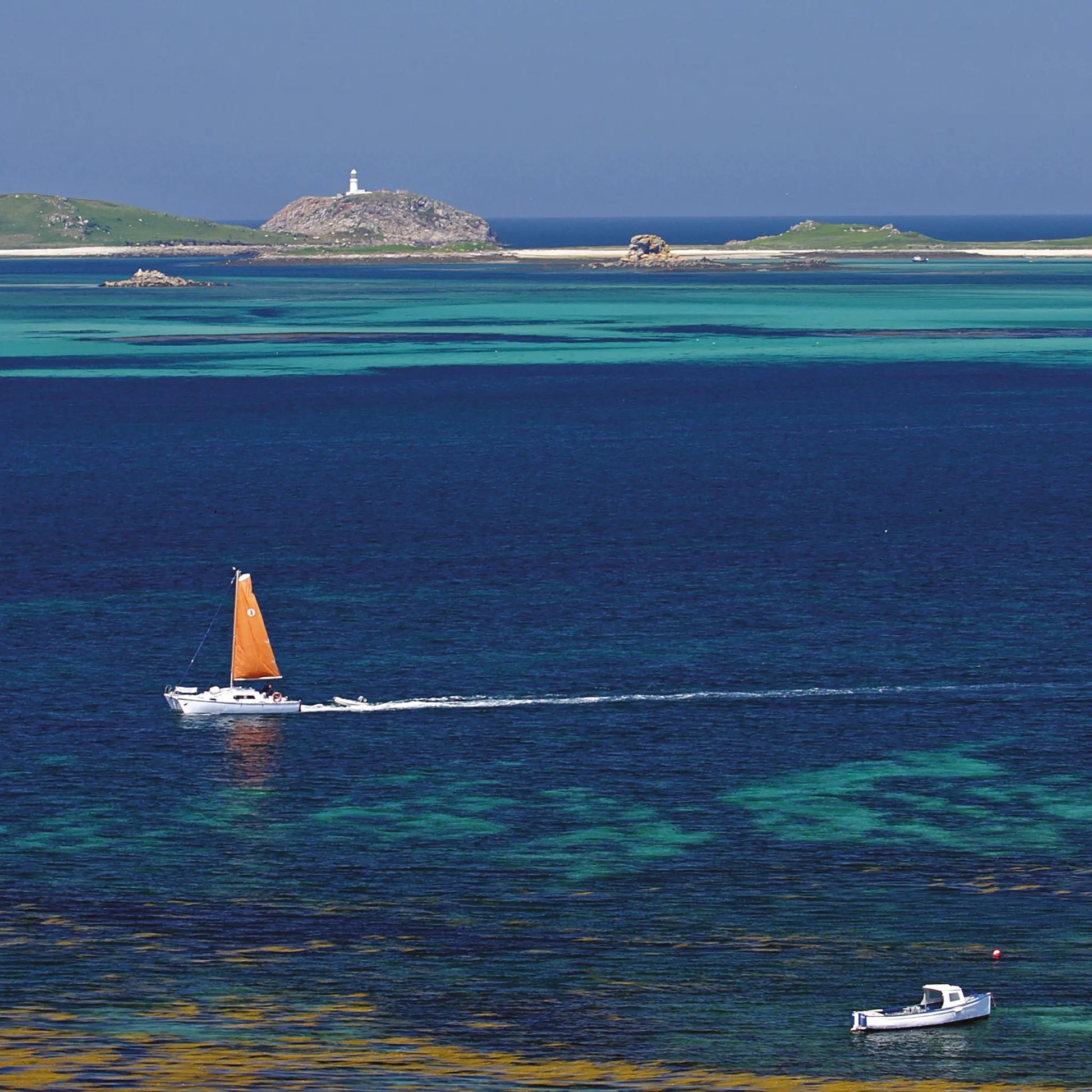 Cornish greetings card image of blue and aquamarine waters in front of Round Island Light, a yacht in the foreground with orange sail