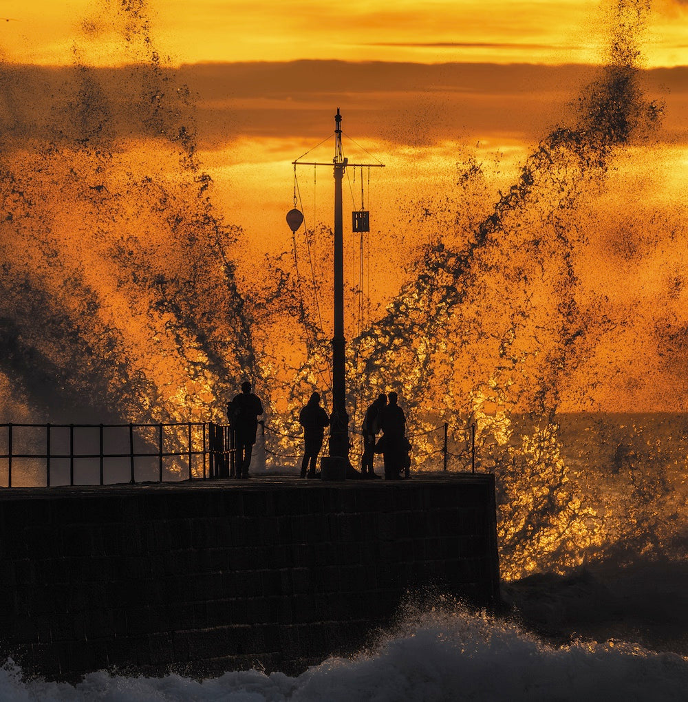 An Evocative Cornwall greetings card depicting people silhouetted at the end of the pier with a large wave crashing in front of them, lit with orange light by the setting sun