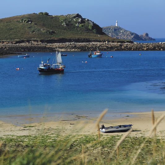 An Evocative Cornwall greetings card showing a blue fishing boat passing others at anchor between two islands, with a small boat resting on the sandy shore and a lighthouse visible in the distance amidst blue sea and sky