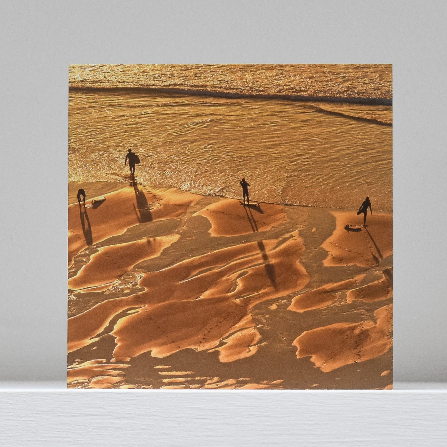 Greetings card on shelf showing image of 4 surfers preparing to enter sea at sunset