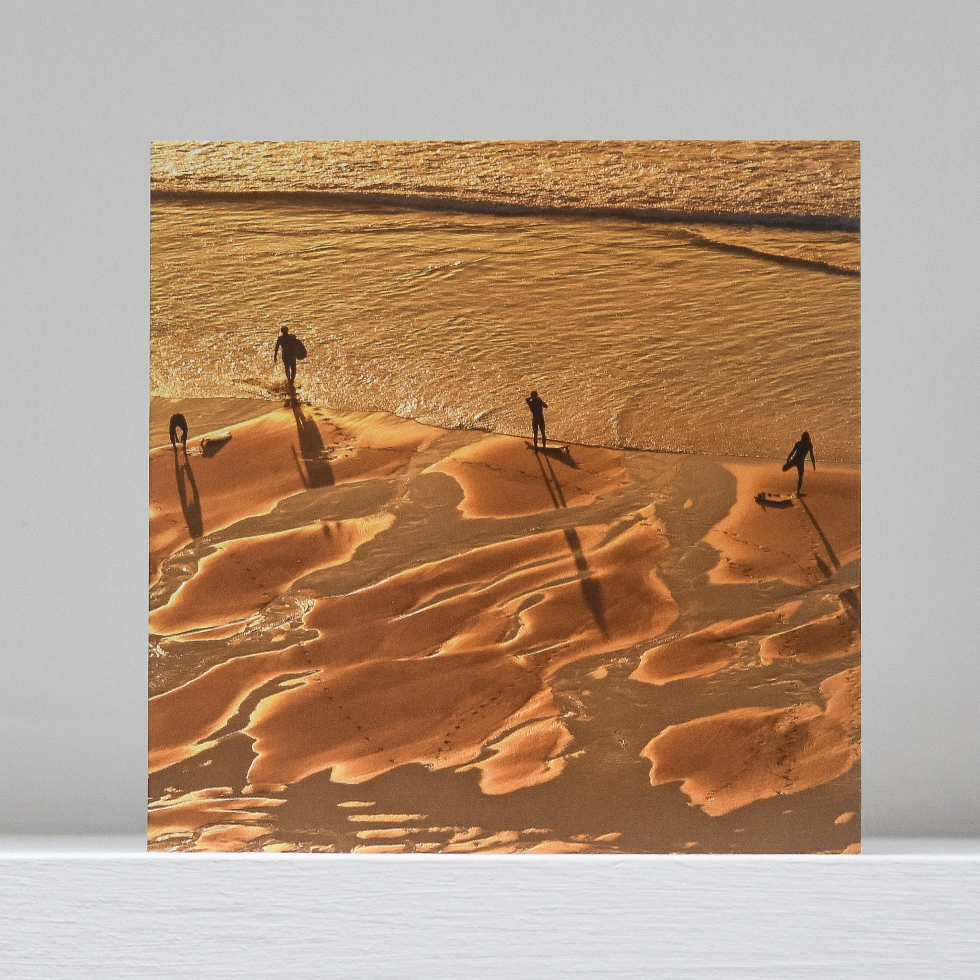 Greetings card on shelf showing image of 4 surfers preparing to enter sea at sunset