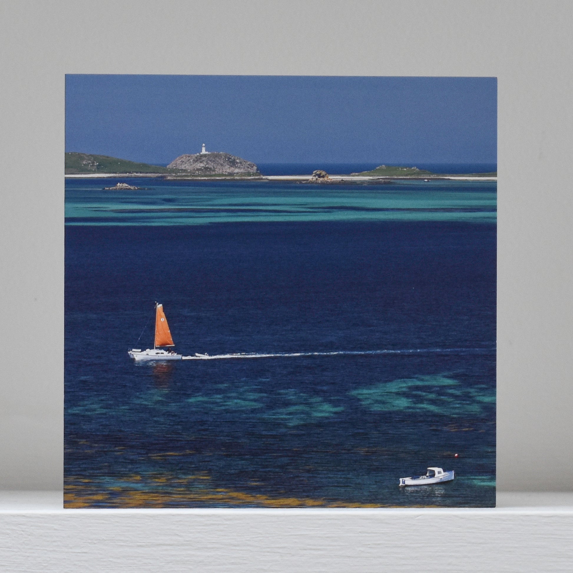 Greetings card on shelf showing image of a catamaran with an orange sail passing Round Island, with lines of blue and turquoise visible in the sea