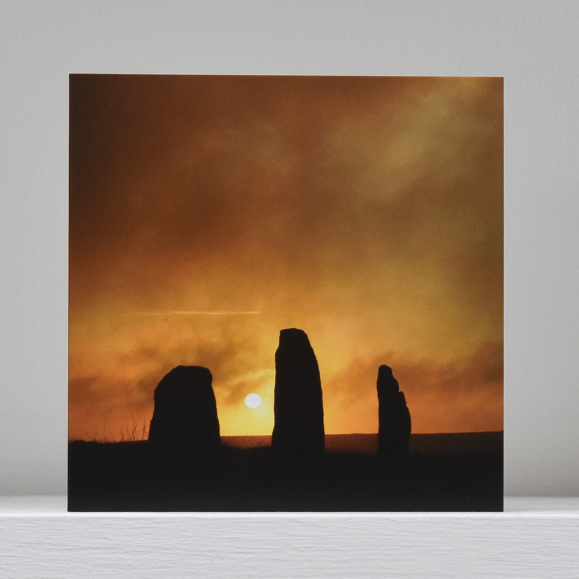 Greetings card on shelf showing image of three stones within the Nine Maidens stone circle in silhouette, with the setting sun behind them and the trail of a plane visible in the orange sky