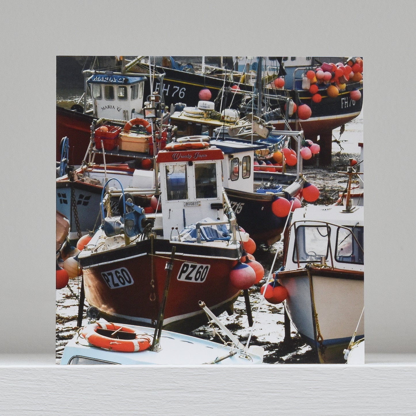 Greetings card on shelf showing image of fishing boats in Mevagissey harbour at low tide - a jumble of colourful boats and buoys.