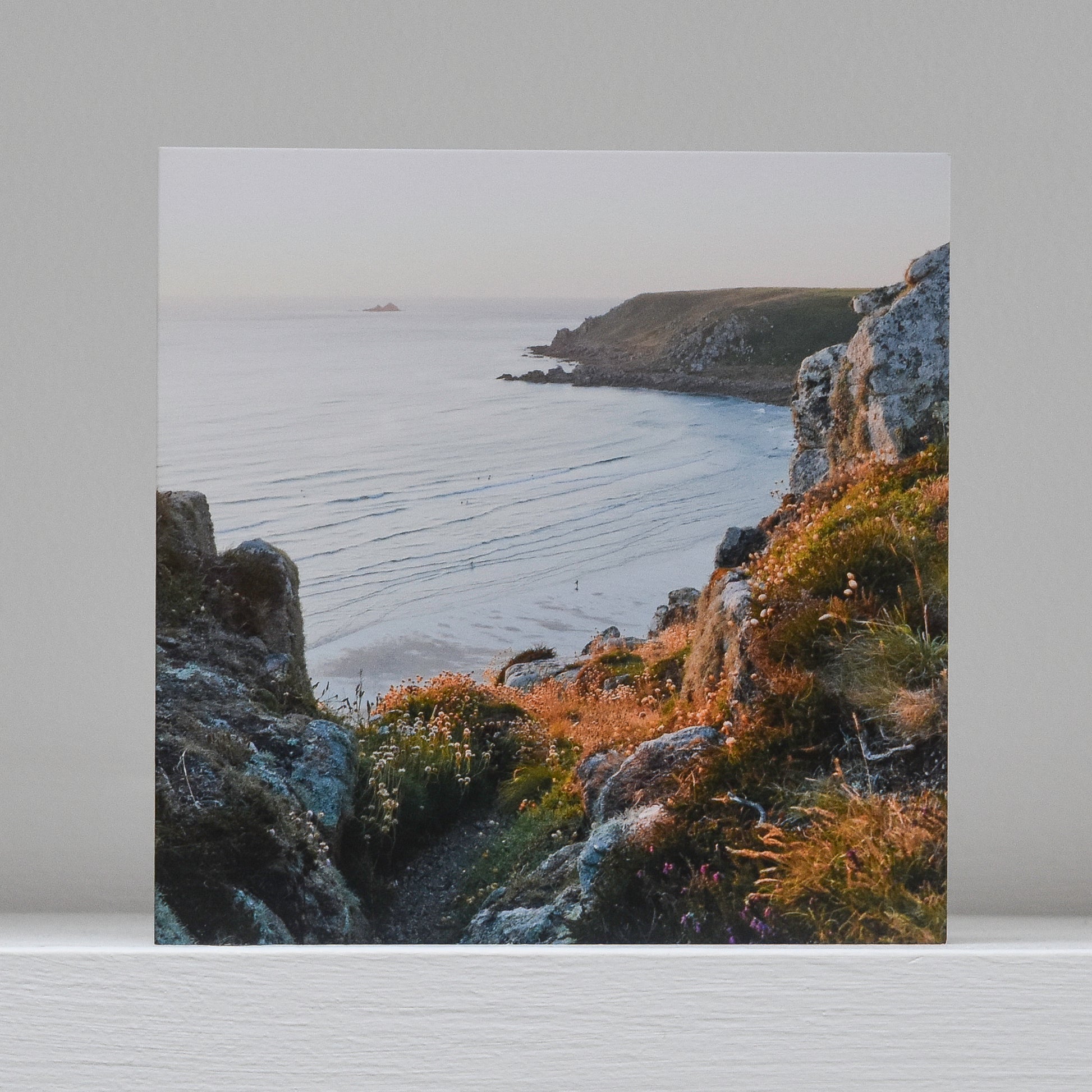 Greetings card on shelf showing image of distant surfer on the beach at Gwynver near Land's End, looking down from the top of the cliffs between granite boulders and wildflowers