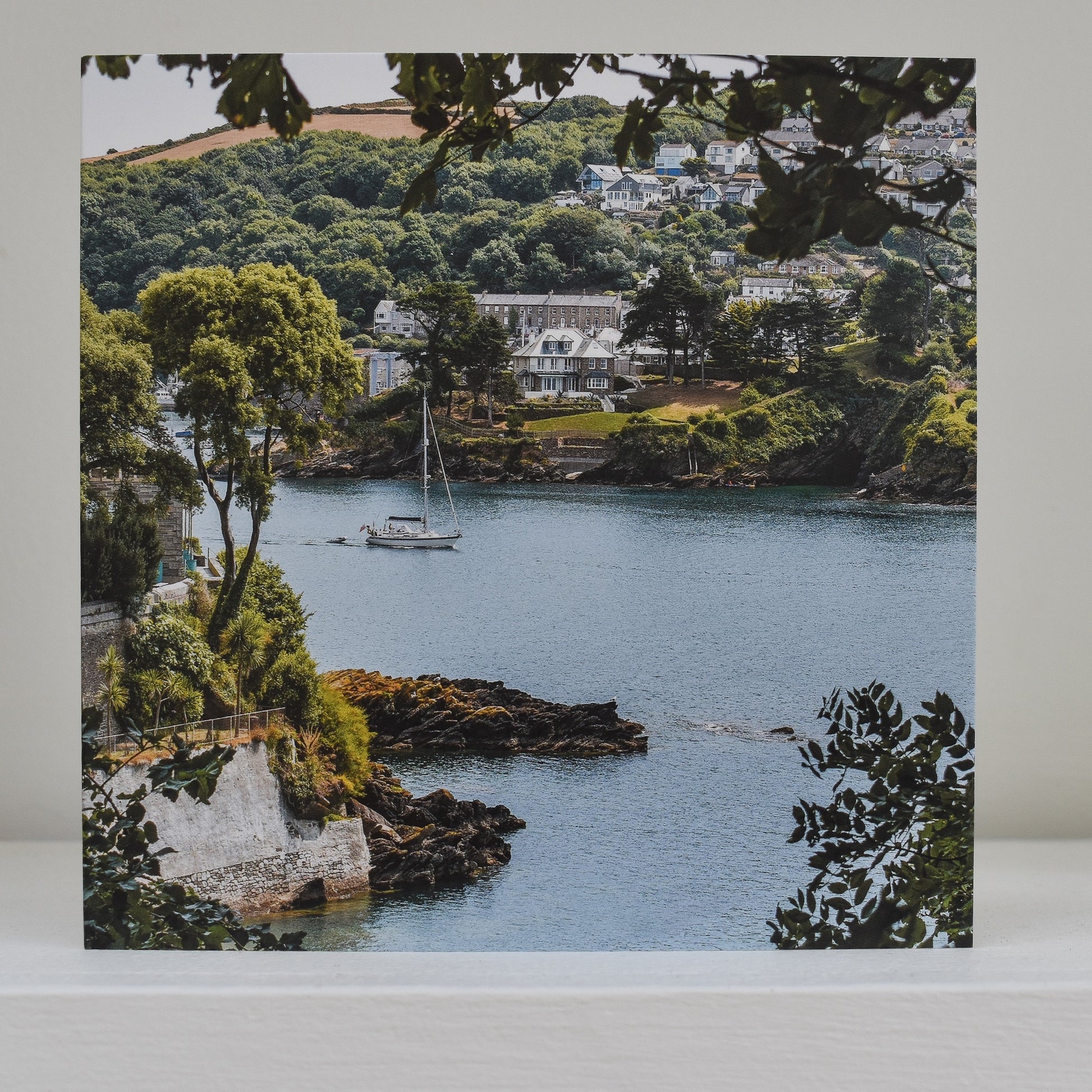 Greeting card on shelf, showing an image from Readymoney Cove, Fowey looking towards Polruan with a yacht sailing out to sea