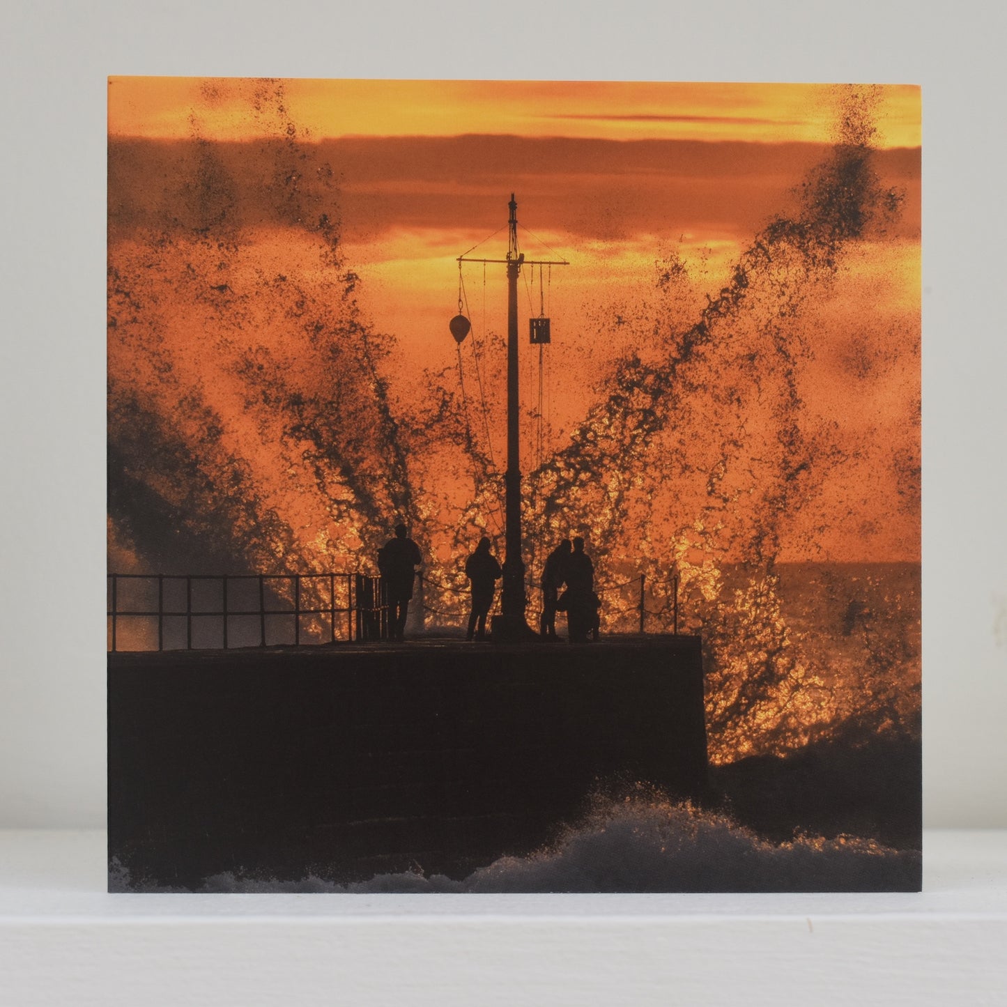 Greeting card on shelf showing image by Geoff Squibb of the pier at Porthleven almost swamped by an enormous wave, which is backlit by the setting sun