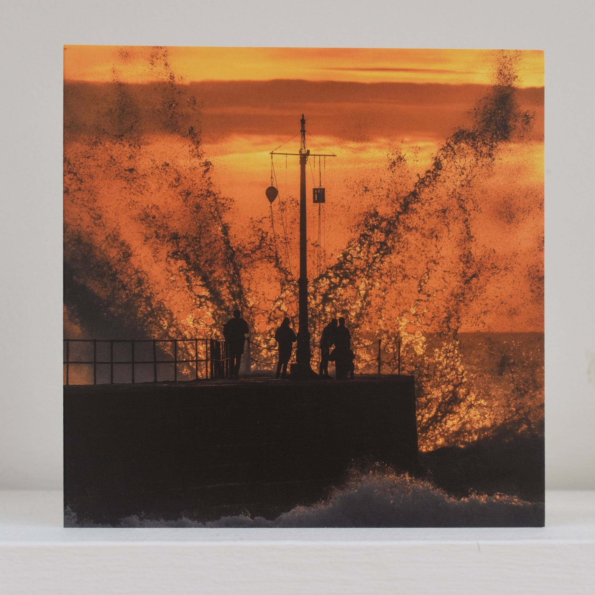 Greeting card on shelf showing image by Geoff Squibb of the pier at Porthleven almost swamped by an enormous wave, which is backlit by the setting sun