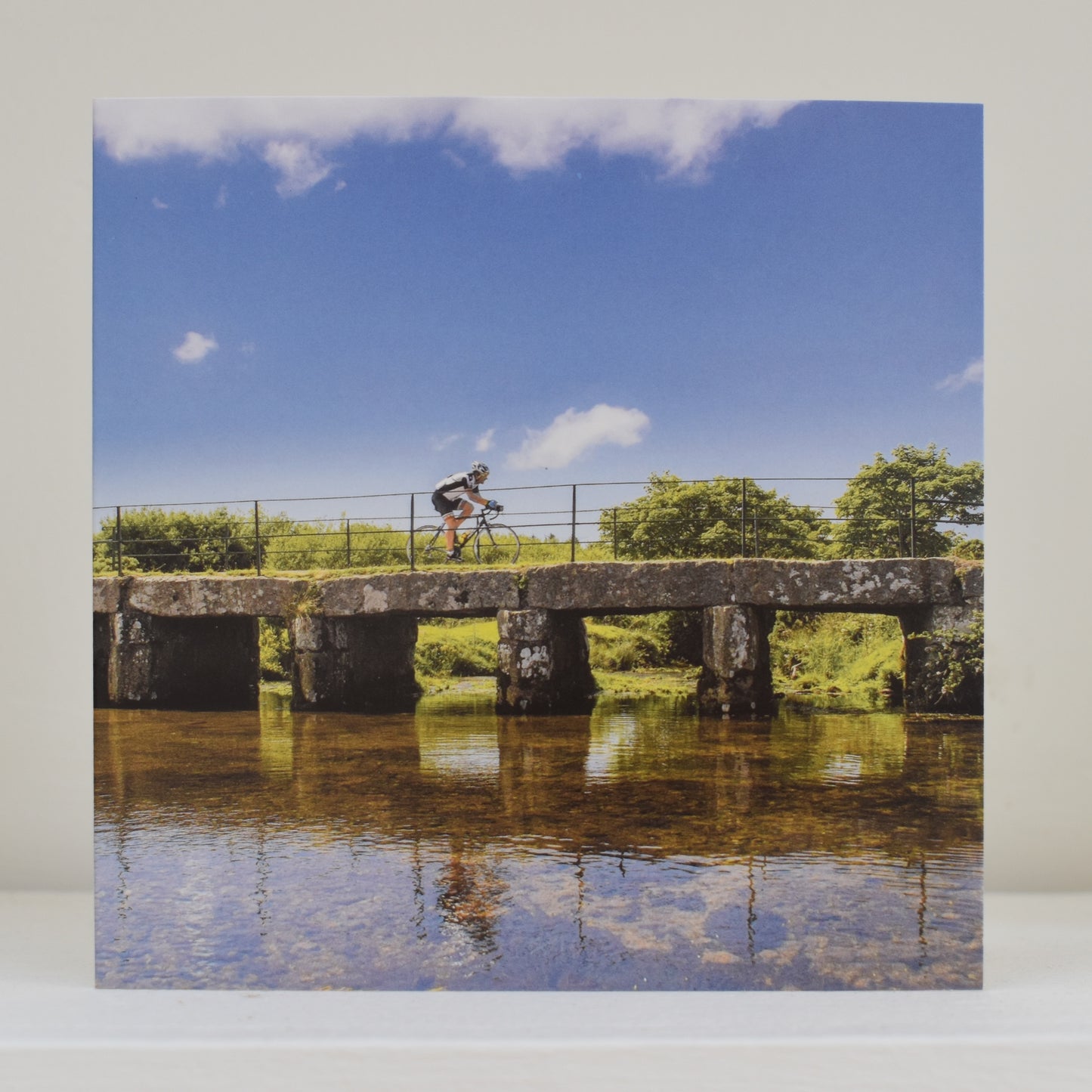 Greeting card on shelf, with an image by Janie Smith showing a cyclist crossing the clapper bridge at Delank on Bodmin Moor