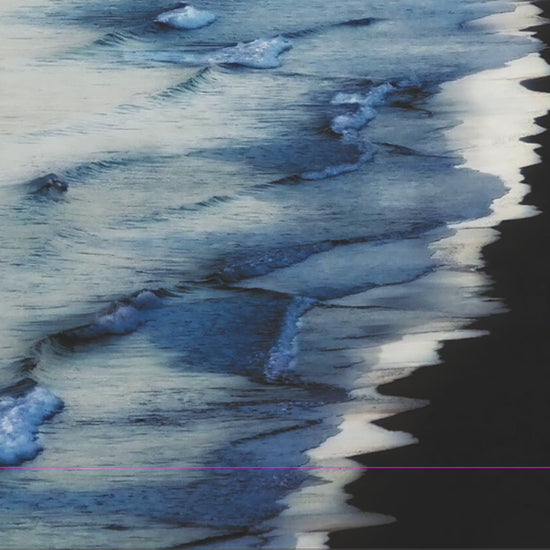 Panning up over the Indigo Shore, white and inky blue waves lap on a dark shore