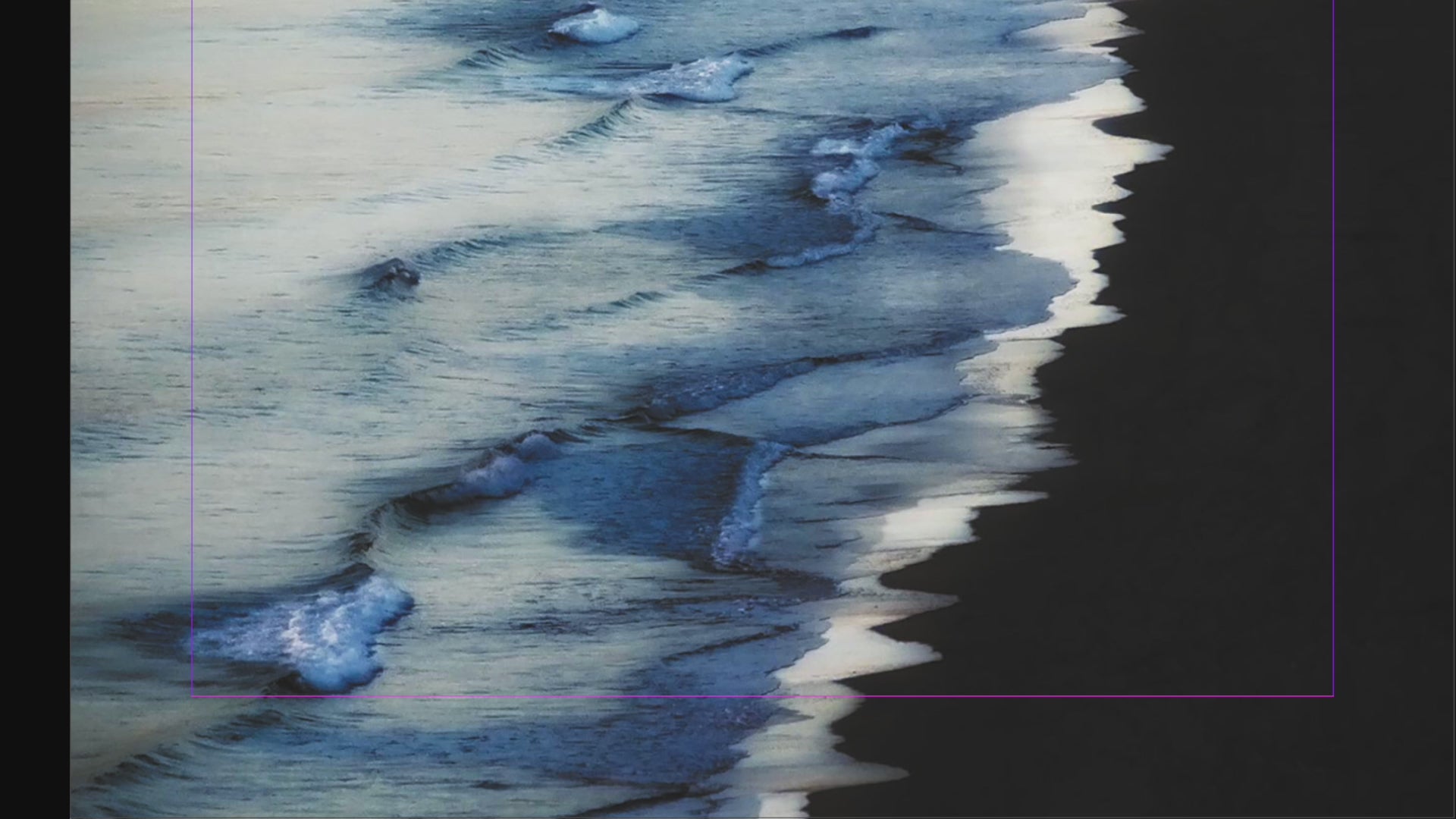 Panning up over the Indigo Shore, white and inky blue waves lap on a dark shore