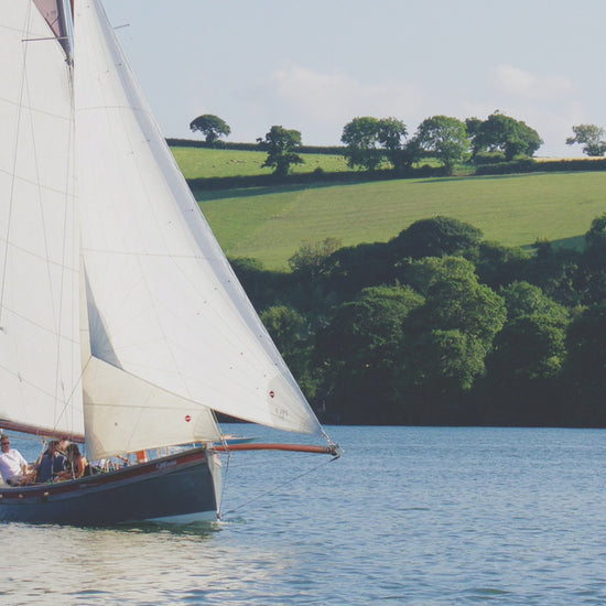 Video animation of a traditional Falmouth Working Boat competing at a local regatta, with Sunbeams also visible racing in the background.  The blue of water and sky, with the green fields and trees of the far shore.
