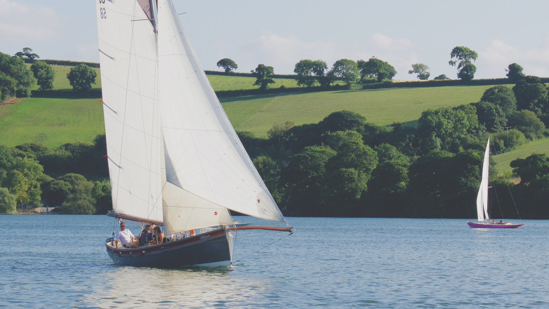Video animation of a traditional Falmouth Working Boat competing at a local regatta, with Sunbeams also visible racing in the background.  The blue of water and sky, with the green fields and trees of the far shore.